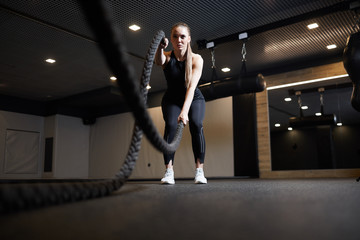 Fototapeta na wymiar Full length wide angle portrait of focused young woman doing battle rope exercises during cross workout in dark gym, copy space