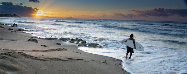 Lone surfer at a Hawaiian shore about to enter the water at sunrise