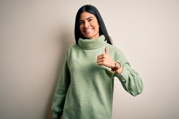 Young beautiful hispanic woman wearing green winter sweater over isolated background doing happy thumbs up gesture with hand. Approving expression looking at the camera showing success.