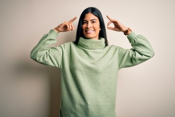 Young beautiful hispanic woman wearing green winter sweater over isolated background smiling pointing to head with both hands finger, great idea or thought, good memory