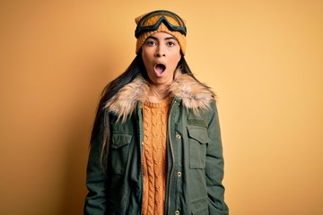 Young beautiful hispanic woman wearing ski glasses and coat for winter weather afraid and shocked with surprise and amazed expression, fear and excited face.