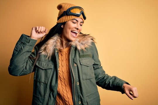 Young beautiful hispanic woman wearing ski glasses and coat for winter weather Dancing happy and cheerful, smiling moving casual and confident listening to music