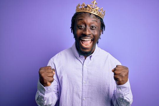 Young african american man wearing golden crown of king over isolated purple background celebrating surprised and amazed for success with arms raised and open eyes. Winner concept.