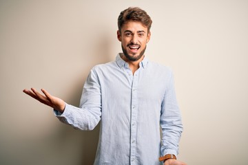 Young handsome man with beard wearing striped shirt standing over white background smiling cheerful with open arms as friendly welcome, positive and confident greetings