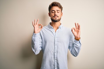 Young handsome man with beard wearing striped shirt standing over white background relaxed and smiling with eyes closed doing meditation gesture with fingers. Yoga concept.