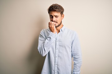 Young handsome man with beard wearing striped shirt standing over white background looking stressed and nervous with hands on mouth biting nails. Anxiety problem.