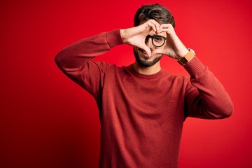 Young handsome man with beard wearing glasses and sweater standing over red background Doing heart shape with hand and fingers smiling looking through sign