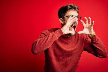 Young handsome man with beard wearing glasses and sweater standing over red background Shouting angry out loud with hands over mouth