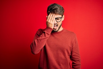 Young handsome man with beard wearing glasses and sweater standing over red background Yawning tired covering half face, eye and mouth with hand. Face hurts in pain.
