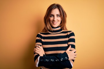 Young beautiful brunette woman wearing striped turtleneck sweater over yellow background happy face smiling with crossed arms looking at the camera. Positive person.