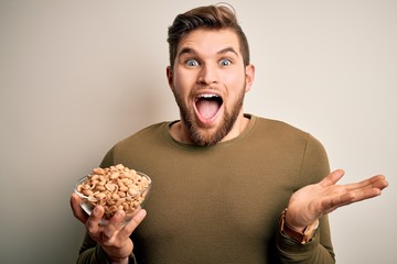 Young blond man with beard and blue eyes holding bowl with healthy salty peanuts very happy and excited, winner expression celebrating victory screaming with big smile and raised hands