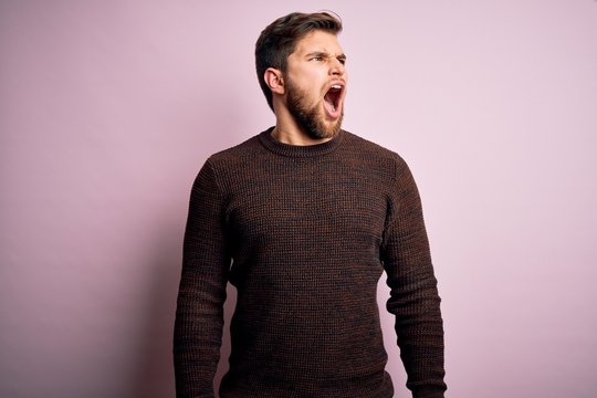 Young blond man with beard and blue eyes wearing casual sweater over pink background angry and mad screaming frustrated and furious, shouting with anger. Rage and aggressive concept.