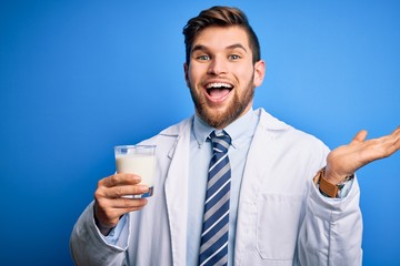 Young blond doctor man with beard and blue eyes wearing coat drinking glass of milk very happy and excited, winner expression celebrating victory screaming with big smile and raised hands