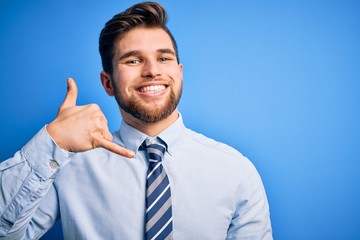 Young blond businessman with beard and blue eyes wearing elegant shirt and tie standing smiling doing phone gesture with hand and fingers like talking on the telephone. Communicating concepts.