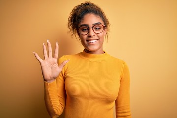 Young beautiful african american girl wearing sweater and glasses over yellow background showing and pointing up with fingers number five while smiling confident and happy.