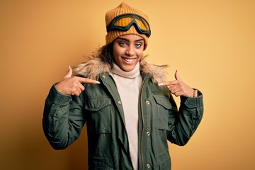 Young african american afro skier girl wearing snow sportswear and ski goggles looking confident with smile on face, pointing oneself with fingers proud and happy.