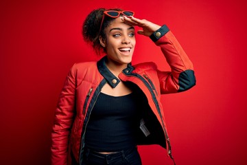 Beautiful african american girl wearing red jacket and sunglasses over isolated background very happy and smiling looking far away with hand over head. Searching concept.