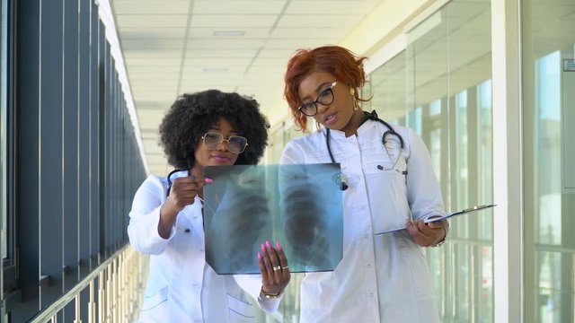 Two female african-american doctors examines x-ray of lungs, holding it in hands indoors. Two specialists holds transparent image of chest in arms, and carefully researches it