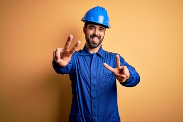 Mechanic man with beard wearing blue uniform and safety helmet over yellow background smiling with tongue out showing fingers of both hands doing victory sign. Number two.
