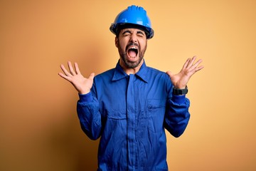 Mechanic man with beard wearing blue uniform and safety helmet over yellow background celebrating...