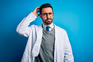 Young handsome doctor man with beard wearing coat and glasses over blue background confuse and wonder about question. Uncertain with doubt, thinking with hand on head. Pensive concept.