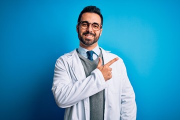 Young handsome doctor man with beard wearing coat and glasses over blue background cheerful with a smile of face pointing with hand and finger up to the side with happy and natural expression on face
