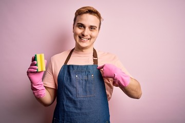 Handsome redhead man doing housework wearing apron and gloves using cleaner scourer pointing finger to one self smiling happy and proud