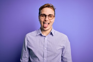 Young handsome redhead man wearing casual shirt and glasses over purple background sticking tongue out happy with funny expression. Emotion concept.
