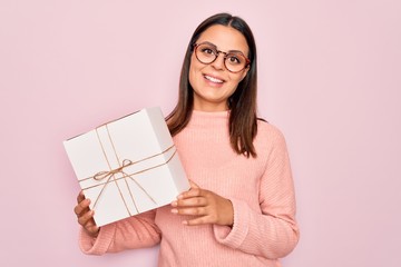 Young beautiful brunette woman holding birthday gift over isolated pink background looking positive and happy standing and smiling with a confident smile showing teeth