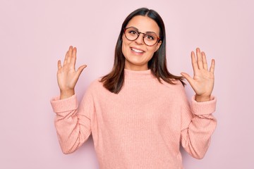 Young beautiful brunette woman wearing casual sweater and glasses over pink background showing and pointing up with fingers number ten while smiling confident and happy.