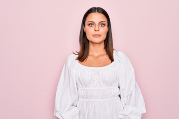 Young beautiful brunette woman wearing casual white dress standing over pink background with serious expression on face. Simple and natural looking at the camera.