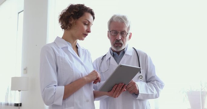Medical team of two generations doctors having teamwork discussion using digital tablet. Old and young professional clinicians staff wearing white coats talking about healthcare doing online research - Powered by Adobe