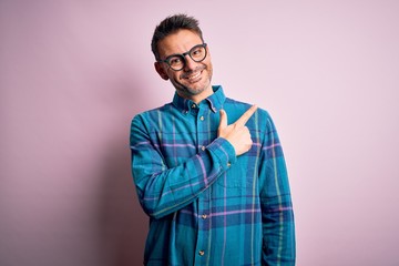 Young handsome man wearing casual shirt and glasses standing over isolated pink background cheerful with a smile of face pointing with hand and finger up to the side with happy and natural expression