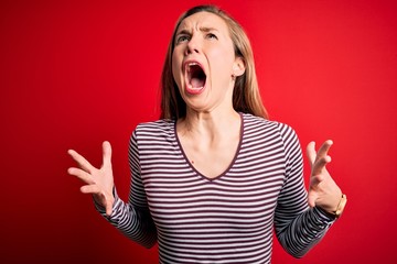 Young beautiful blonde woman wearing casual striped t-shirt over isolated red background crazy and mad shouting and yelling with aggressive expression and arms raised. Frustration concept.