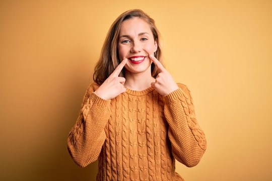 Young beautiful blonde woman wearing casual sweater standing over yellow background Smiling with open mouth, fingers pointing and forcing cheerful smile