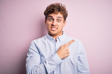 Young blond handsome man with curly hair wearing striped shirt over white background Pointing aside worried and nervous with forefinger, concerned and surprised expression