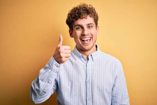 Young blond handsome man with curly hair wearing striped shirt over yellow background doing happy thumbs up gesture with hand. Approving expression looking at the camera showing success.