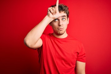 Young blond handsome man with curly hair wearing casual t-shirt over red background making fun of people with fingers on forehead doing loser gesture mocking and insulting.