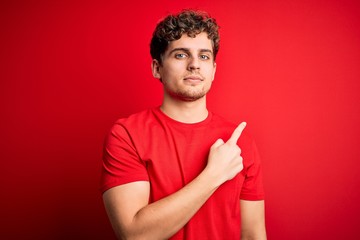 Young blond handsome man with curly hair wearing casual t-shirt over red background Pointing with hand finger to the side showing advertisement, serious and calm face