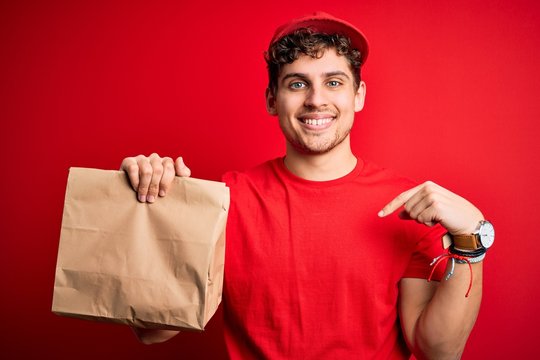 Young blond delivery man with curly hair wearing cap holding paper bag with food with surprise face pointing finger to himself