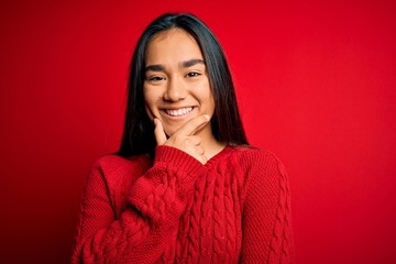 Fototapeta premium Young beautiful asian woman wearing casual sweater standing over isolated red background looking confident at the camera smiling with crossed arms and hand raised on chin. Thinking positive.