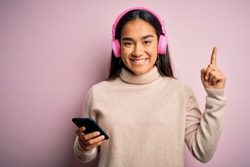 Young beautiful asian woman listening to music using smartphone and headphones surprised with an...