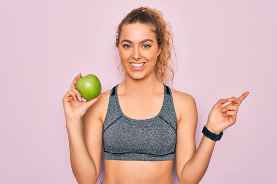 Young beautiful blonde sporty woman with blue eyes holding healthy green apple fruit very happy pointing with hand and finger to the side