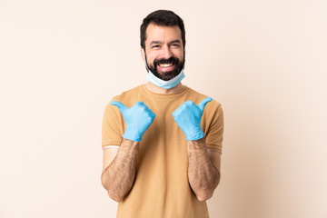 Fototapeta premium Caucasian man with beard protecting from the coronavirus with a mask and gloves over isolated background with thumbs up gesture and smiling