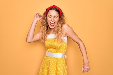 Beautiful blonde pin-up woman with blue eyes wearing diadem standing over yellow background Dancing happy and cheerful, smiling moving casual and confident listening to music