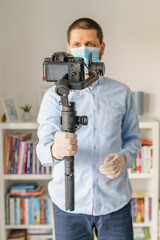 Front view on caucasian adult man blogger standing in at home n day holding gimbal stabilizer with camera making video about pandemic virus disease spread wearing protective mask gloves for prevention
