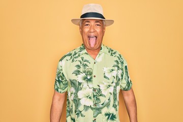 Middle age senior grey-haired man wearing summer hat and floral shirt on beach vacation sticking tongue out happy with funny expression. Emotion concept.