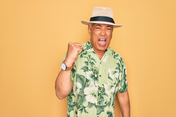 Middle age senior grey-haired man wearing summer hat and floral shirt on beach vacation angry and mad raising fist frustrated and furious while shouting with anger. Rage and aggressive concept.