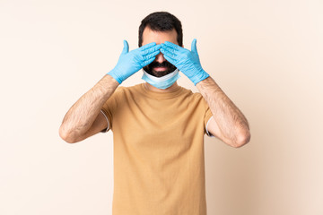 Caucasian man with beard protecting from the coronavirus with a mask and gloves over isolated background covering eyes by hands and smiling