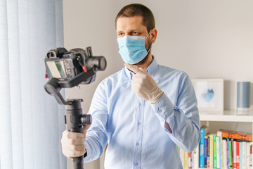 Fototapeta na wymiar Front view on caucasian adult man blogger standing in at home n day holding gimbal stabilizer with camera making video about pandemic virus disease spread wearing protective mask gloves for prevention